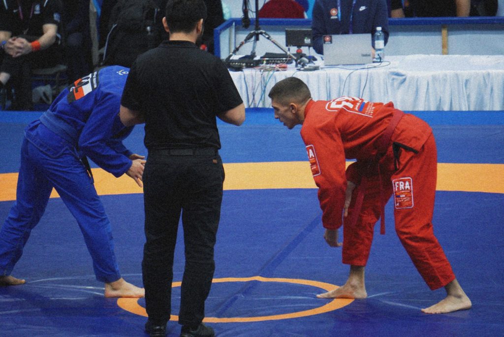 Two Brazilian Jiu-Jitsu competitors in red and blue gis facing each other on the mat, with a referee between them.