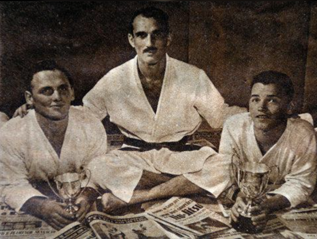 Photographie historique de trois pratiquants de jiu-jitsu brésilien en kimono, dont deux présentent des trophées, avec des journaux sportifs au premier plan.